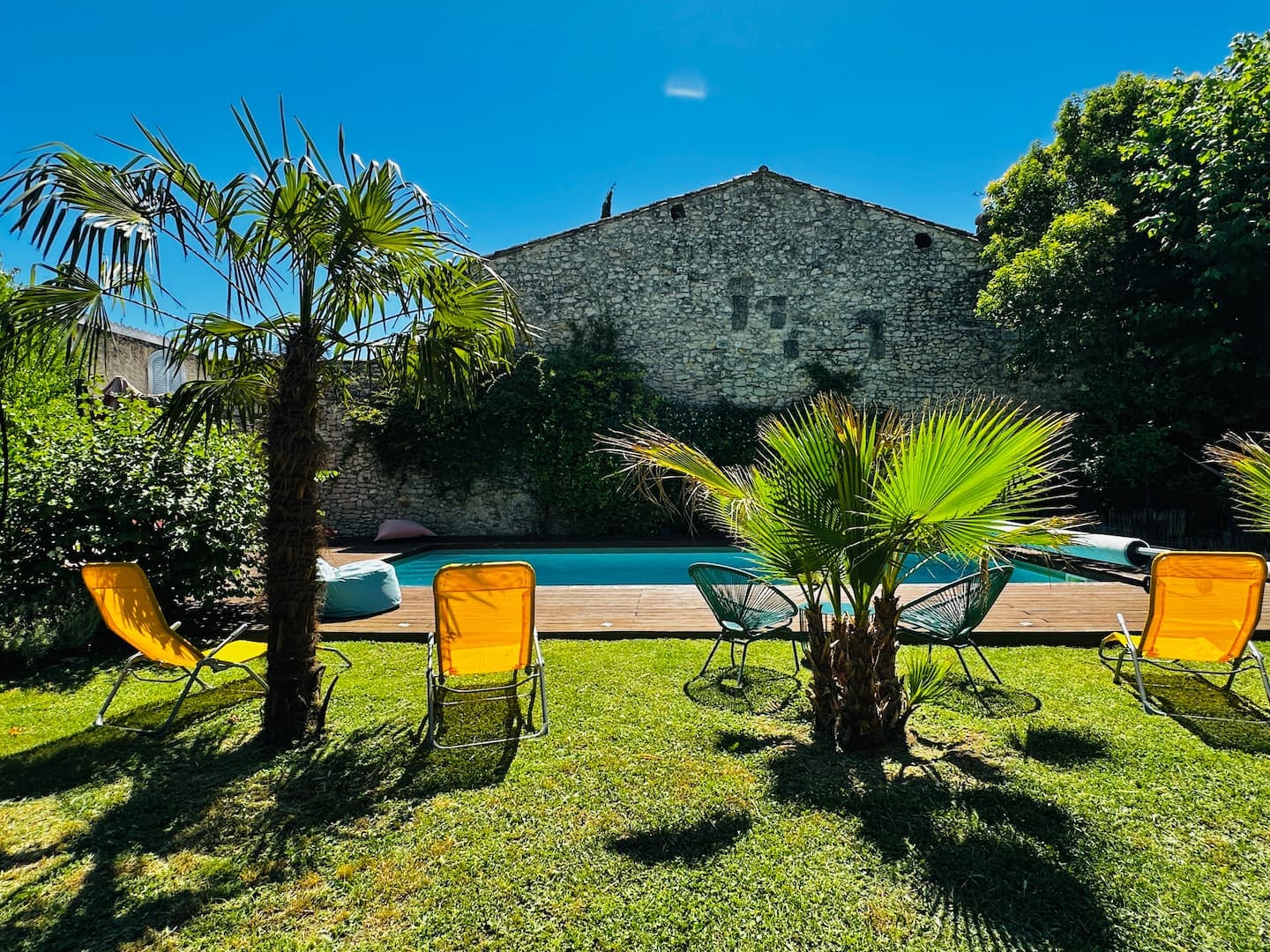 La piscine et la façade en pierre de la Vieille Magnanerie, palmiers et ciel bleu