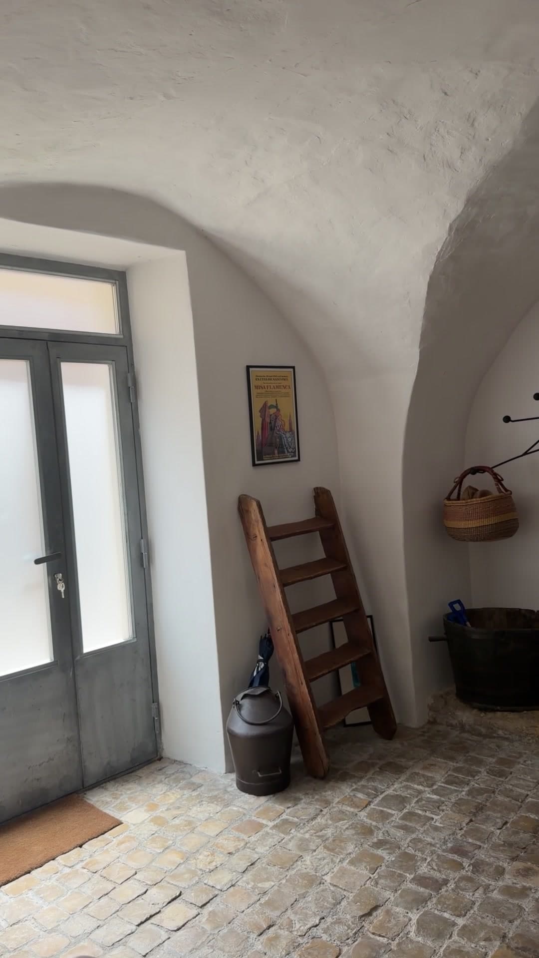 Porte vitrée grise ouverte sur le jour, échelle ancienne et bidon de lait sous la voûte plein cintre